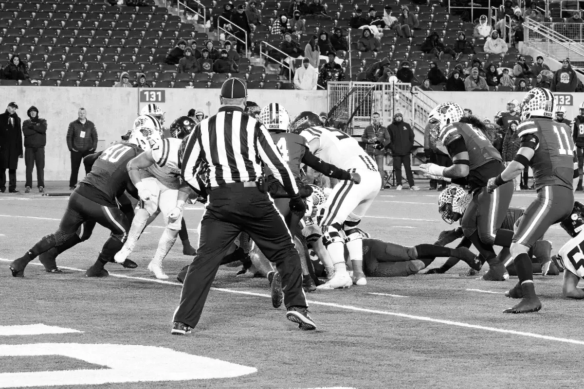 A referee observes a group of football players from two teams scrambling for the ball during a game, with spectators in the stadium seats behind them.