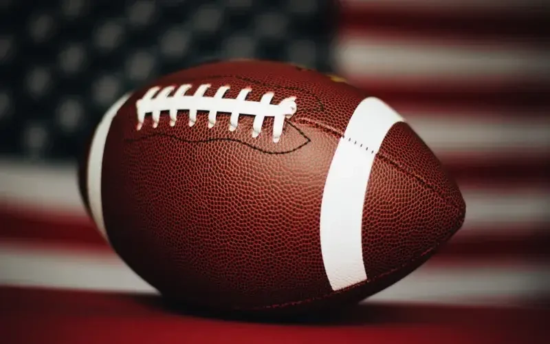 A close-up of an American football with white laces and stripes, positioned in front of a blurred United States flag background.