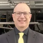 A man wearing glasses, a black suit, and a yellow tie stands smiling in front of an open garage with equipment visible in the background.