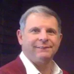 A middle-aged man with short gray hair is smiling, wearing a red sweater over a white shirt, against a dark background.