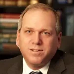 A middle-aged man in a suit and tie sits in an office setting with bookshelves in the background.