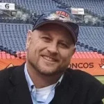 A man in a suit jacket and baseball cap sits in a stadium with empty blue seats and an orange "Broncos" banner visible behind him.