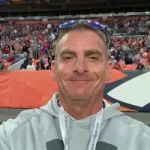 A man wearing a gray hoodie and a lanyard takes a selfie at a crowded outdoor sports stadium.