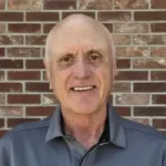 Smiling older man with short gray hair wearing a gray collared shirt, standing in front of a red brick wall.