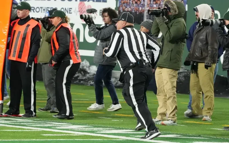 A football referee runs on the field near sideline officials and photographers, all bundled in winter clothing during a game.
