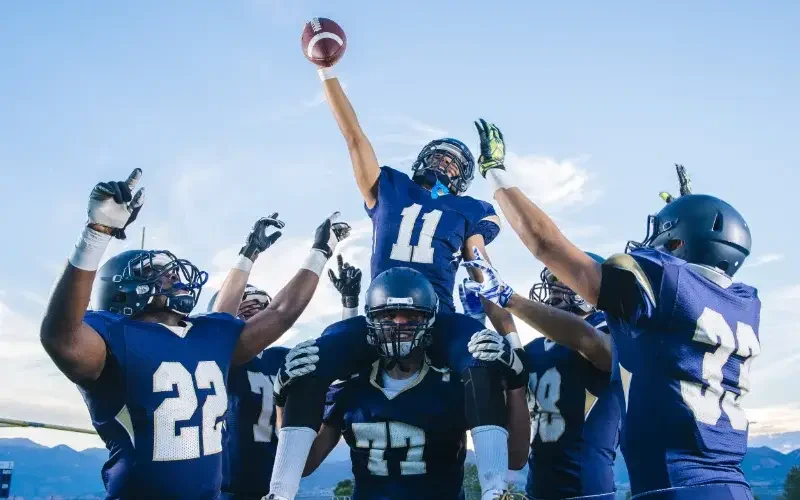 A football player wearing jersey number 11 is lifted by teammates, holding a football above his head in celebration. Other players in blue uniforms cheer around him.
