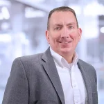 A man in a gray blazer and white shirt smiles while standing indoors with a blurred office background.