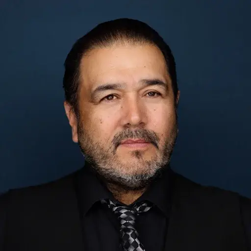 A man with short dark hair, a beard, and a mustache is wearing a black shirt, black vest, and patterned tie, posed against a solid dark blue background.