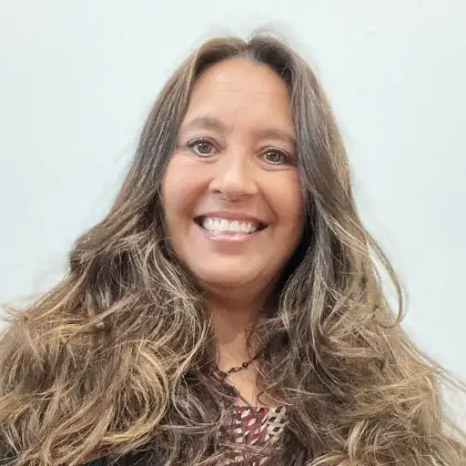 A woman with long, wavy brown hair smiles at the camera. She is wearing a patterned top and a necklace, posing in front of a plain light-colored background.