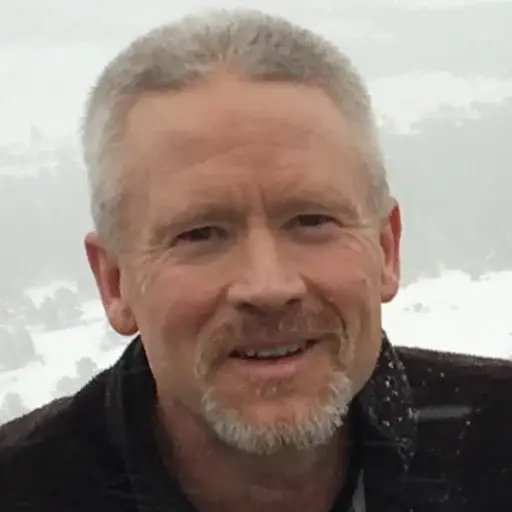 A middle-aged man with short gray hair and a trimmed beard smiles outdoors, wearing a dark jacket. Snowy trees and a cloudy sky appear blurred in the background.
